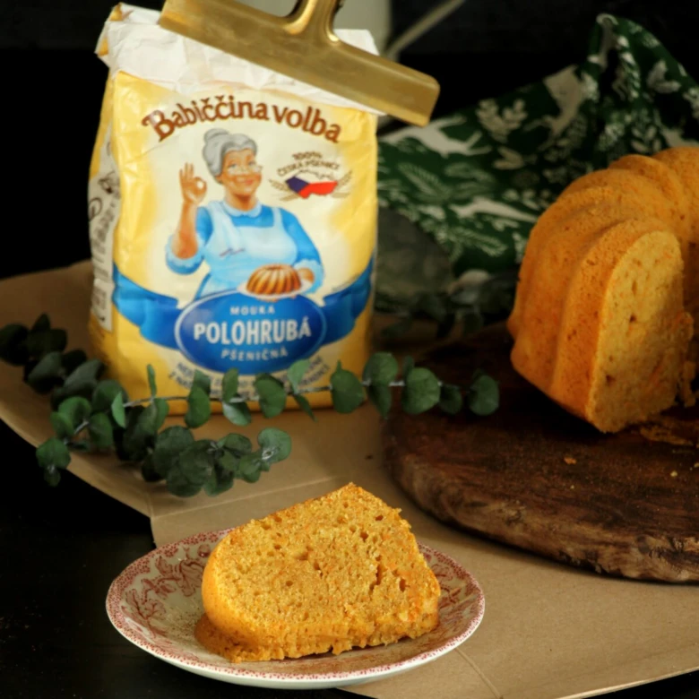Kousek mrkvové bábovky s balíčkem mouky na pozadí.