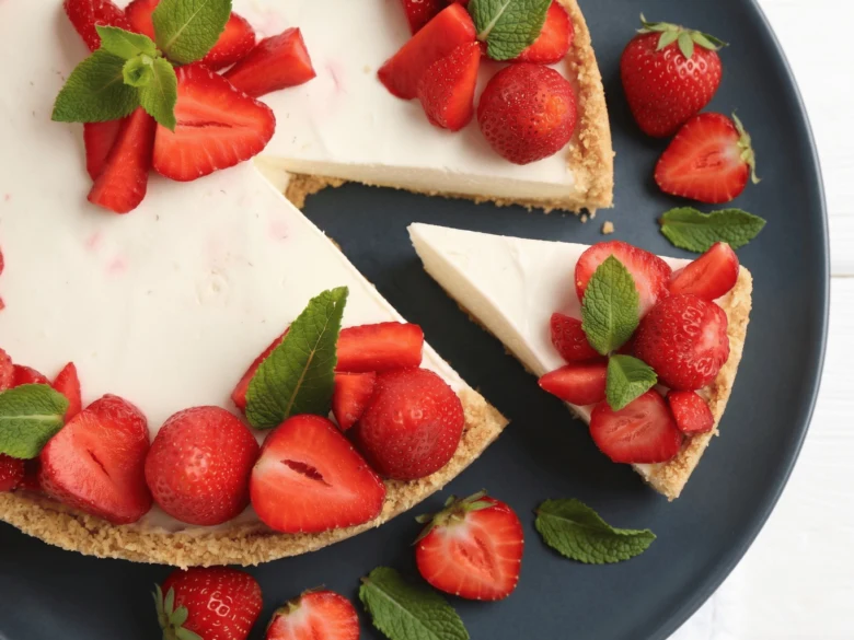 Celý jahodový cheesecake s odseknutým kouskem na tmavém talíři.