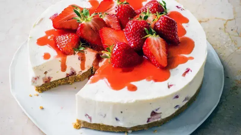 Celý jahodový cheesecake s odseknutým kouskem na bílém talíři.