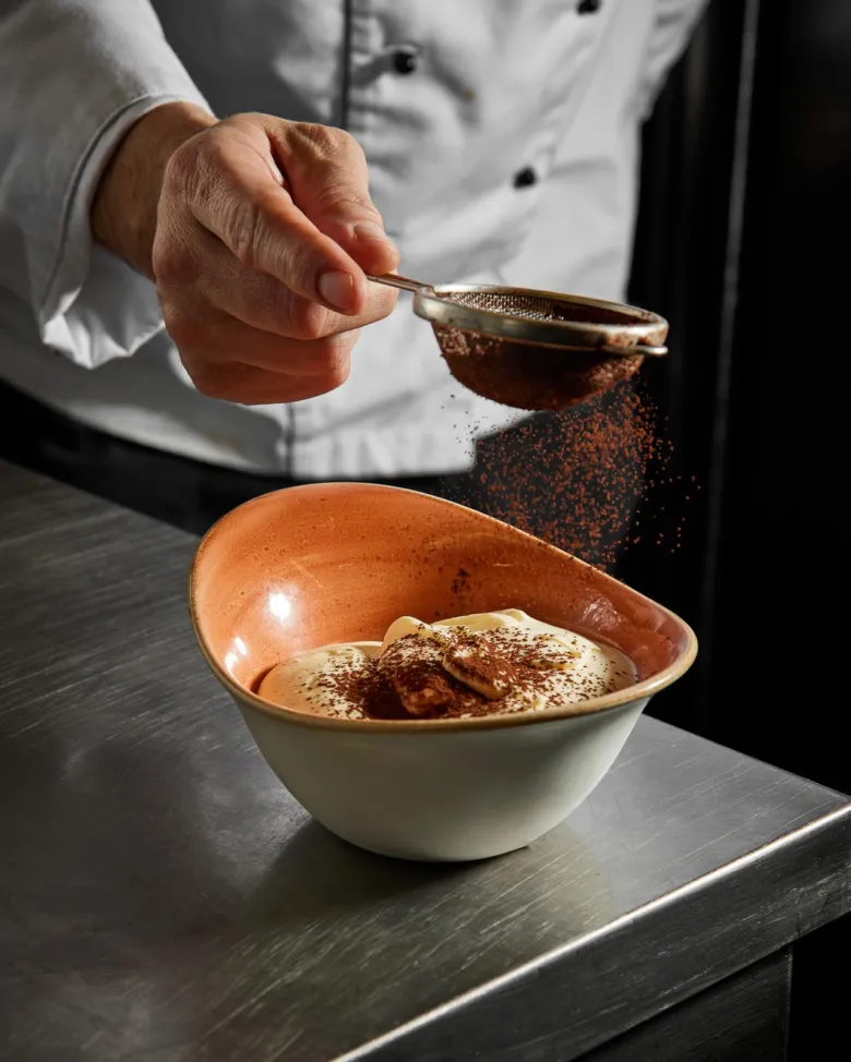 Šéfkuchař posypává čokoládovou moukou italské tiramisu.