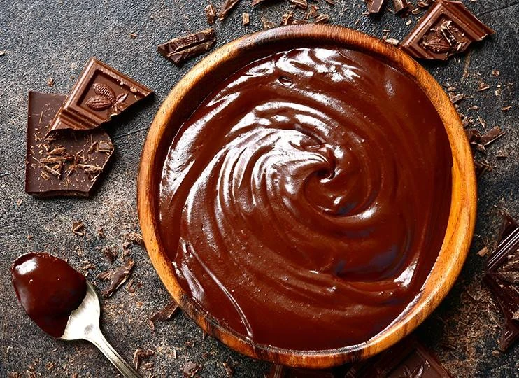 Lesklá čokoládová ganache v dřevěné mísce s kousky tmavé čokolády.