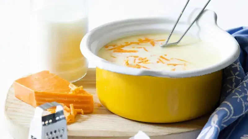 Sýrová omáčka v žlutém hrnci s nastrouhaným sýrem a struhadlem vedle.