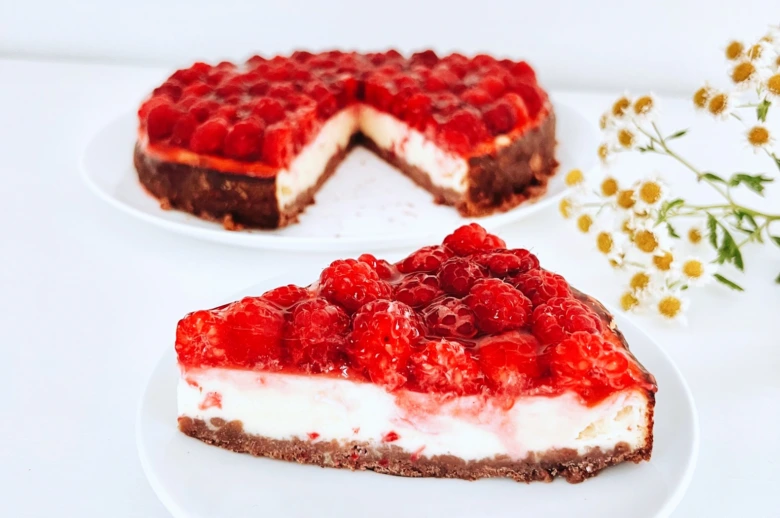 Kus malinového cheesecaku s celým dortem na bílém podkladu.