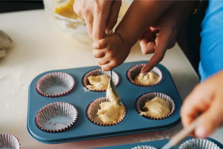 Dospělí s dítětem nalévají těsto do formy na muffiny.
