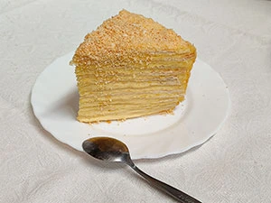 Kousek dortu Napoleon na bílém talíři s lžící.