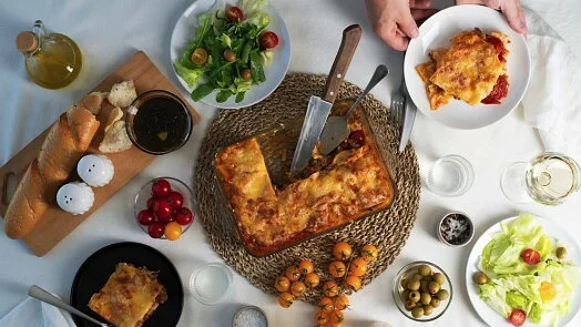 Pohled shora na pečenou lasagnu s saláty, chlebem a zeleninou.
