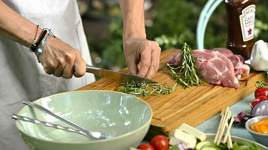 Člověk řezá čerstvé bylinky k masu na dřevěném prknu s ingrediencemi.