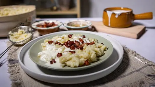 Pohár s haluškami, na kterých je rozlitá bryndza a křupavé uzeniny.