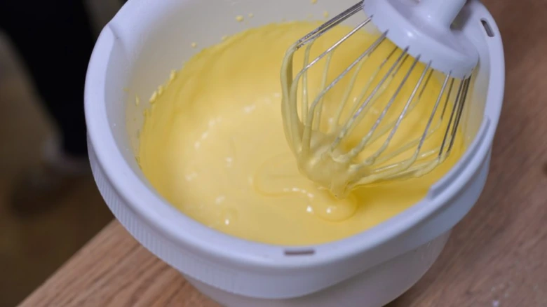 Bílý mixér smíchává žluté těsto v míse na dřevěné desce.