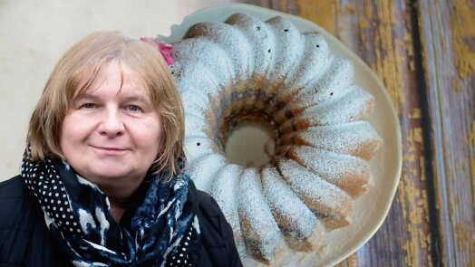 Žena stojí vedle bábovky hrníčkové posypané moučkou.